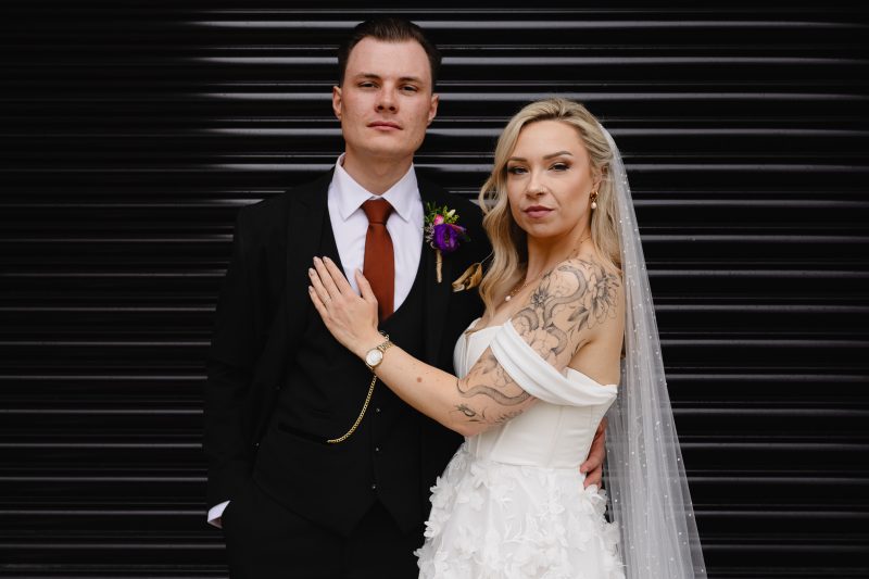 Alternative wedding couple stood together in front of a grey shutter backdrop