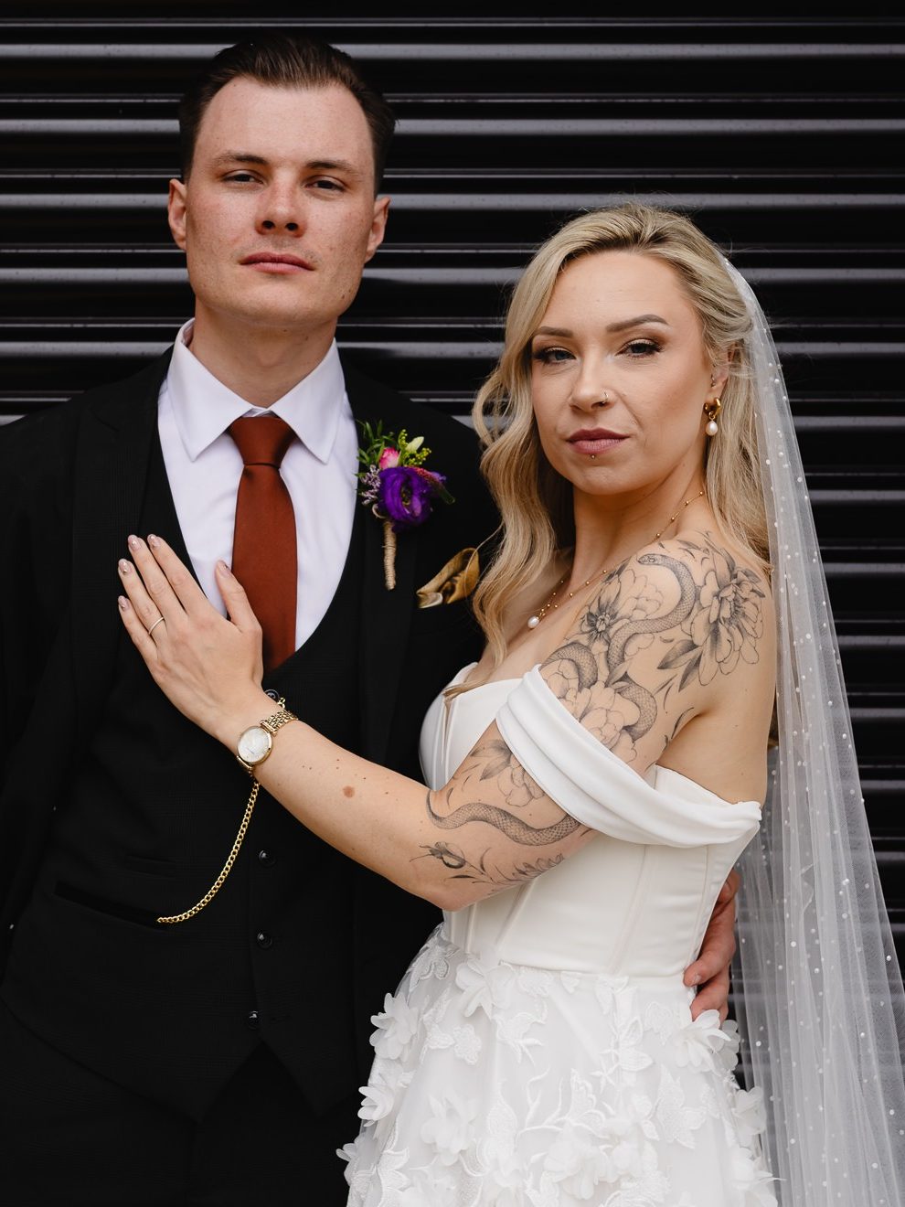 Alternative wedding couple stood together in front of a grey shutter backdrop