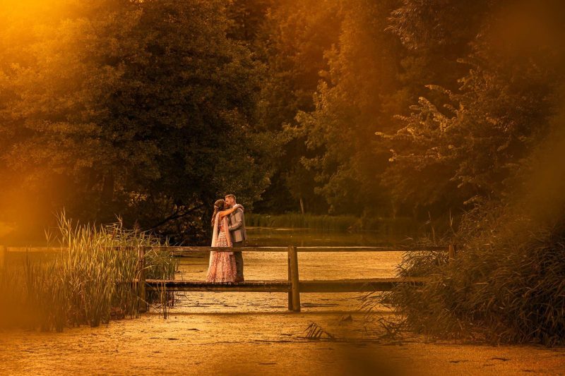 Bride and groom kissing romantically on a bridge over The Great Barn Lake at golden hour sunset in Oxfordshire wearing Indian wedding attire